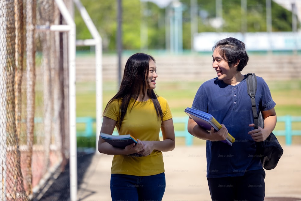 Outside of school, a happy young couple of students stands along a fence, studying a book.