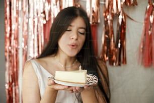 Mulher feliz elegante no vestido elegante soprando vela acesa no pedaço do bolo de aniversário no fundo da guirlanda moderna da borla de ouro rosa no quarto. Comemorando aniversário em casa. Faça um desejo
