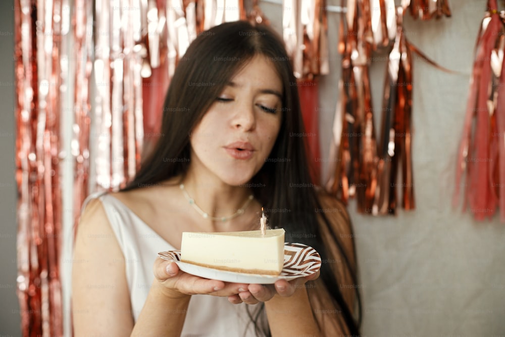 Stylish happy woman in elegant dress blowing burning candle on piece of birthday cake on background of modern rose gold tassel garland in room. Celebrating birthday at home. Make a wish