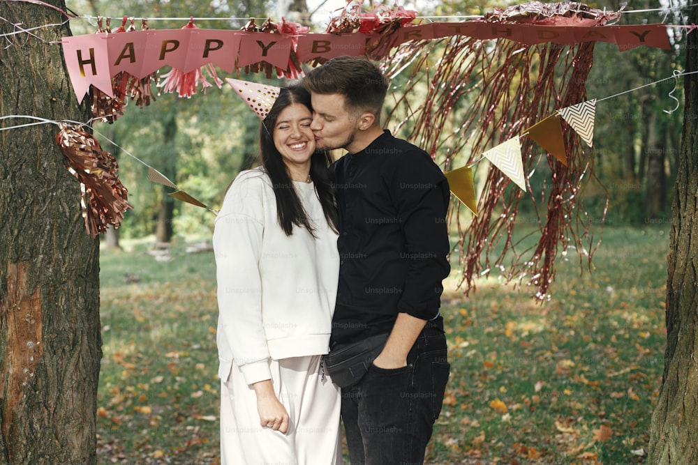 Young stylish couple celebrating birthday with family and friends at picnic party outdoor. Happy woman in party hat and handsome man hugging on background of pink happy birthday garland in park