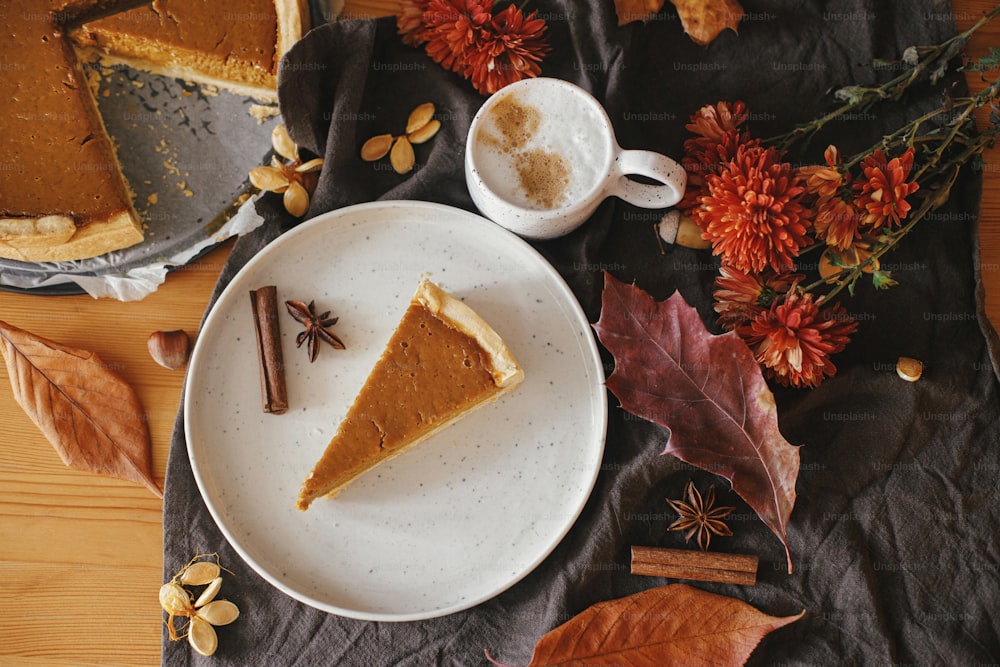 Pumpkin pie slice on modern plate and hot coffee on rustic table with linen napkin, autumn flowers and leaves, anise and cinnamon. Homemade pumpkin tart recipe. Top view. Happy Thanksgiving