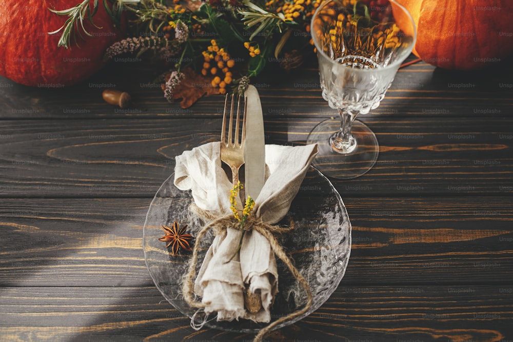 Thanksgiving-Tischdekoration. Moderner Teller mit Vintage-Besteck, Leinenserviette, Kräuter und Glas auf Holztisch mit Kürbissen und Herbstblumenarrangement im Sonnenlicht. Rustikales Catering