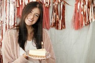 Mulher feliz elegante segurando o pedaço do bolo de aniversário com vela acesa no fundo da guirlanda moderna da borla de ouro rosa no quarto. Comemorando aniversário em casa. Faça um desejo