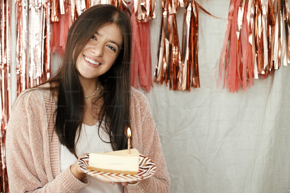 Donna felice alla moda che tiene un pezzo di torta di compleanno con candela accesa sullo sfondo della moderna ghirlanda di nappe in oro rosa in camera. Festeggiare il compleanno a casa. Esprimi un desiderio
