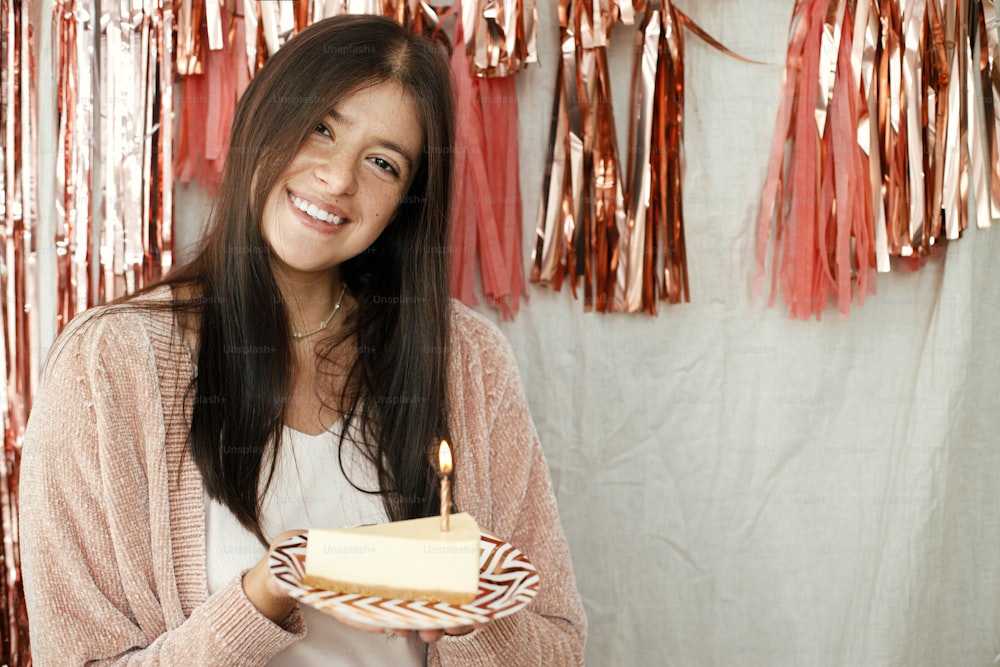 Stylish happy woman holding piece of birthday cake with burning candle on background of modern rose gold tassel garland in room. Celebrating birthday at home. Make a wish