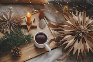 Warm cup of tea with christmas lights, stars, pine trees, candle and pillows on soft bed. Flat lay. Cozy home. Winter hygge. Festive holiday decorations in scandinavian room. Atmospheric  moment