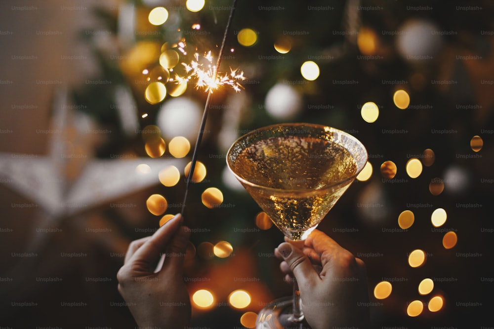 새해 복 많이 받으세요! 크리스마스 트리 조명과 별의 배경에 불타는 폭죽과 샴페인 잔을 들고 있는 손. 불꽃놀이 벵골 빛과 여자 손에 음료. 대기 순간