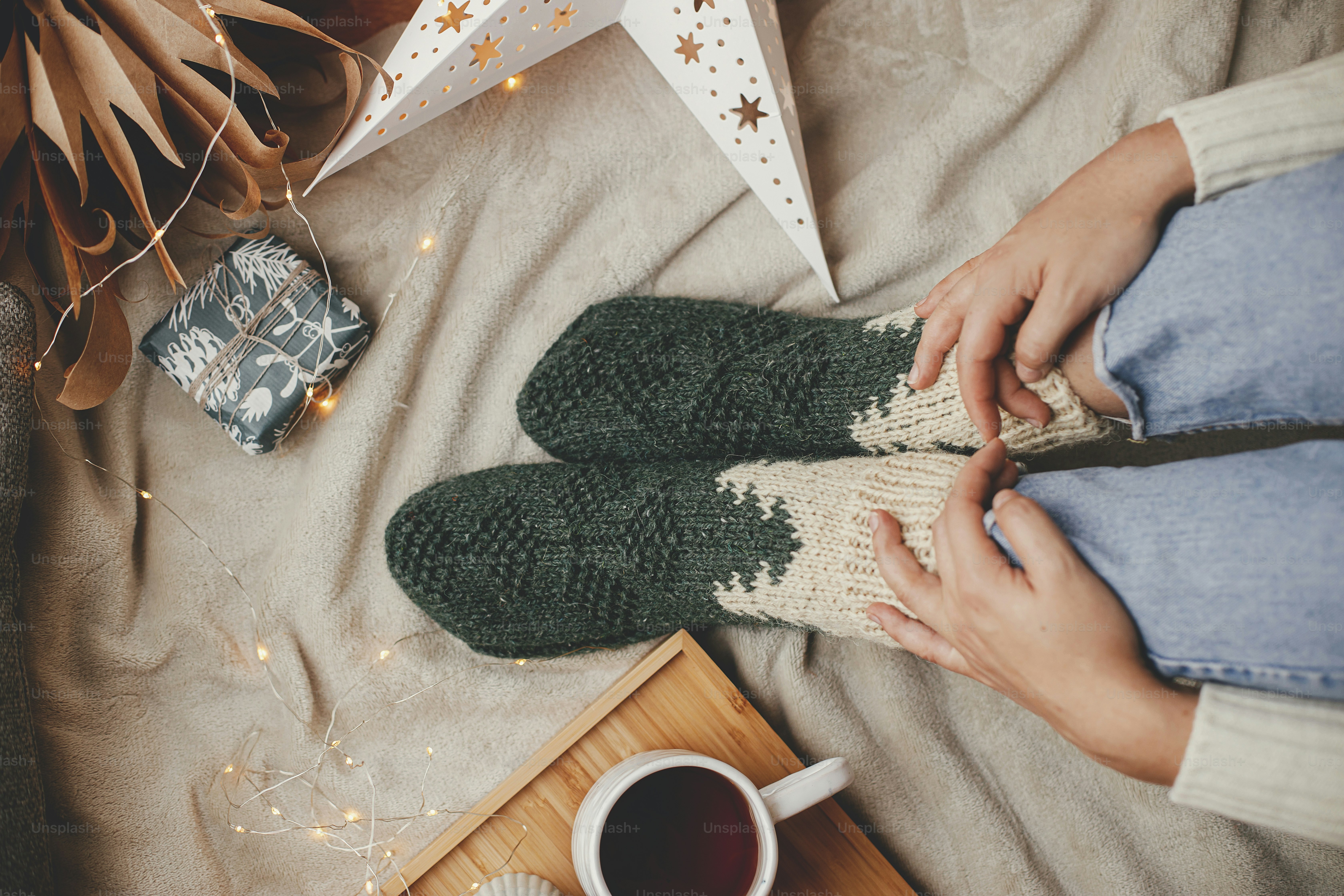 Woman legs in warm socks on soft blanket with cup of tea, christmas stars, golden lights, trees, candle and pillows, top view. Cozy relaxing moments at home. Winter and autumn holidays.