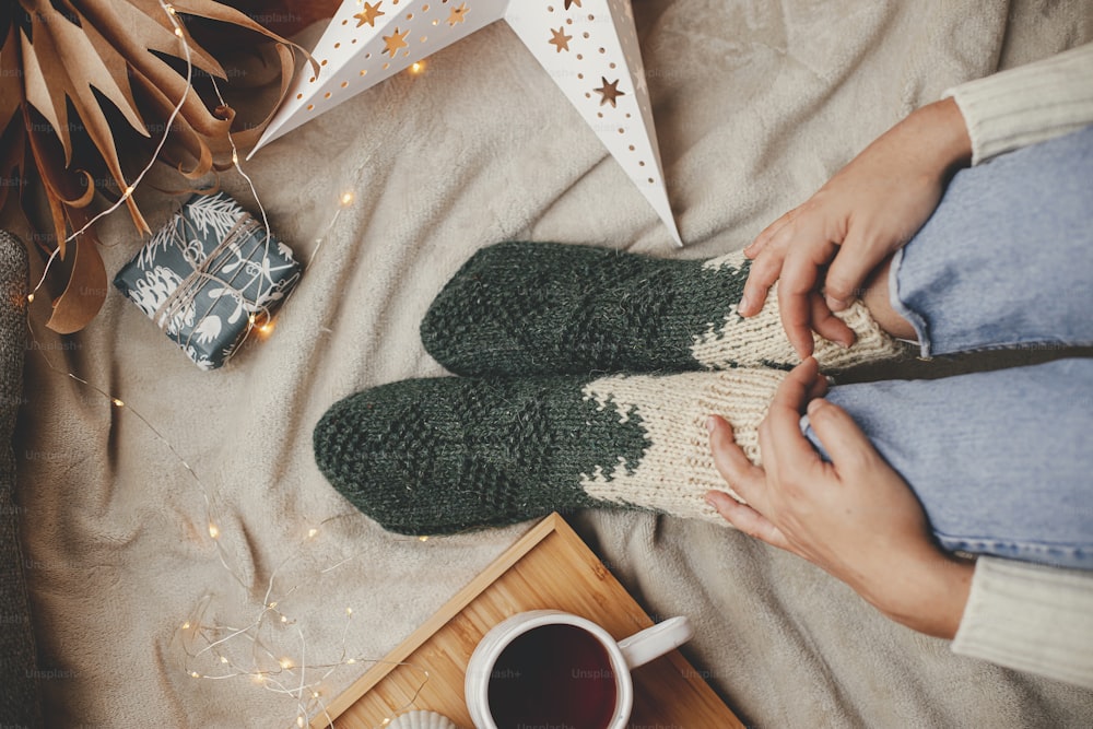 Frauenbeine in warmen Socken auf weicher Decke mit Tasse Tee, Weihnachtssterne, goldene Lichter, Bäume, Kerze und Kissen, Draufsicht. Gemütliche Entspannungsmomente zu Hause. Winter- und Herbstferien.