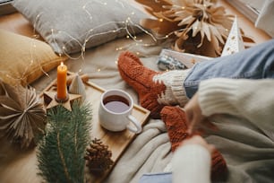 Woman legs in warm socks on soft blanket with cup of tea, christmas stars, golden lights, trees, candle and pillows. Cozy relaxing moments at home. Winter and autumn holidays.