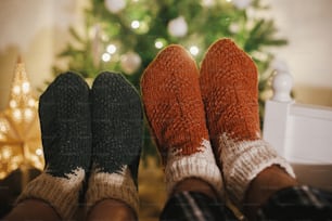 Couples feet in cozy woolen socks on background of christmas tree in lights in festive evening room. Celebrating winter holidays together, cozy family moments at home. Stylish warm socks