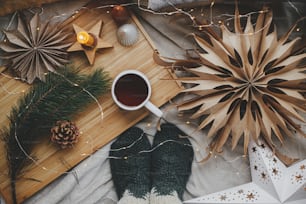 Woman feet in woolen socks on soft bed with warm cup of tea, christmas stars, golden lights, trees, candle and pillows. Top view. Cozy moments at scandinavian home. Winter and autumn holidays