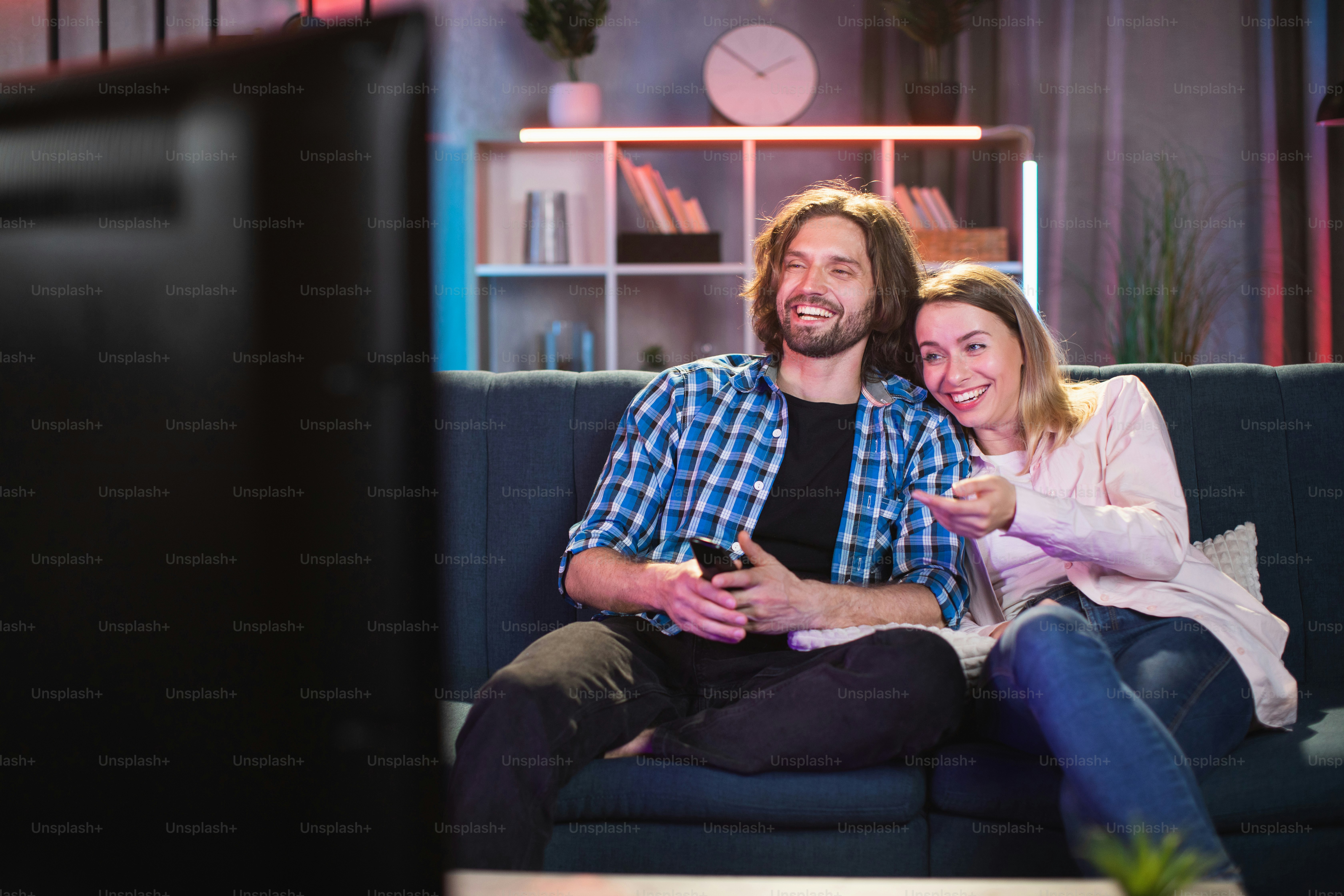 Caucasian young man and woman smiling sincerely while watching comedy on TV during evening time. Happy couple enjoying free time at home.
