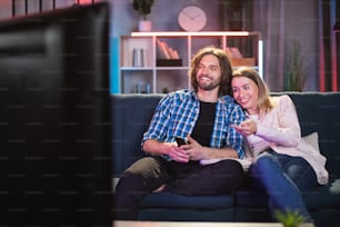 Caucasian young man and woman smiling sincerely while watching comedy on TV during evening time. Happy couple enjoying free time at home.