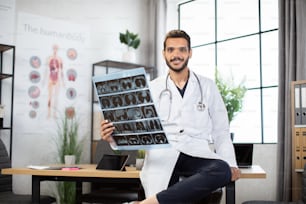 Medicine, radiology and healthcare concept. Happy smiling male indian doctor white coat, holding MRI x-ray scan image and looking at camera over medical office at hospital background