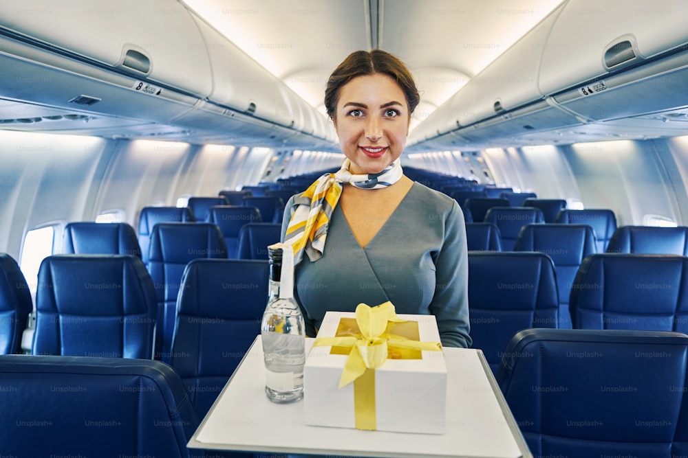 Ritratto in su di un'assistente di volo sorridente e adorabile che trasporta una scatola regalo e una bottiglia di bevanda per i passeggeri