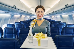 Retrato de cintura para arriba de una azafata encantadora sonriente que lleva una caja de regalos y una botella de bebida para los pasajeros