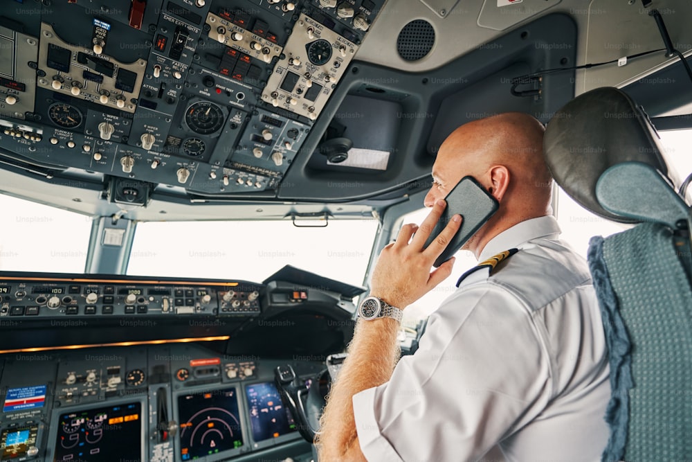 Rückansicht eines professionellen kaukasischen Flugkapitäns mit seinem Smartphone im Cockpit