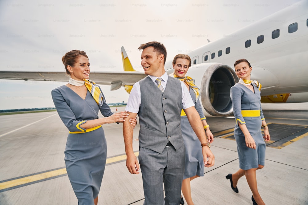 Joyous male flight attendant accompanied by three elegant cute young Caucasian air hostesses in uniforms