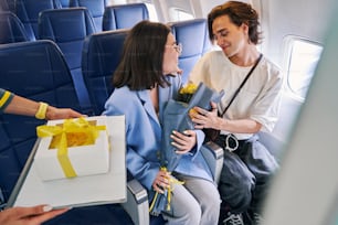 Joven de cabello oscuro con un ramo de hermosas flores y su novio atractivo complacido sentado en la cabina del avión