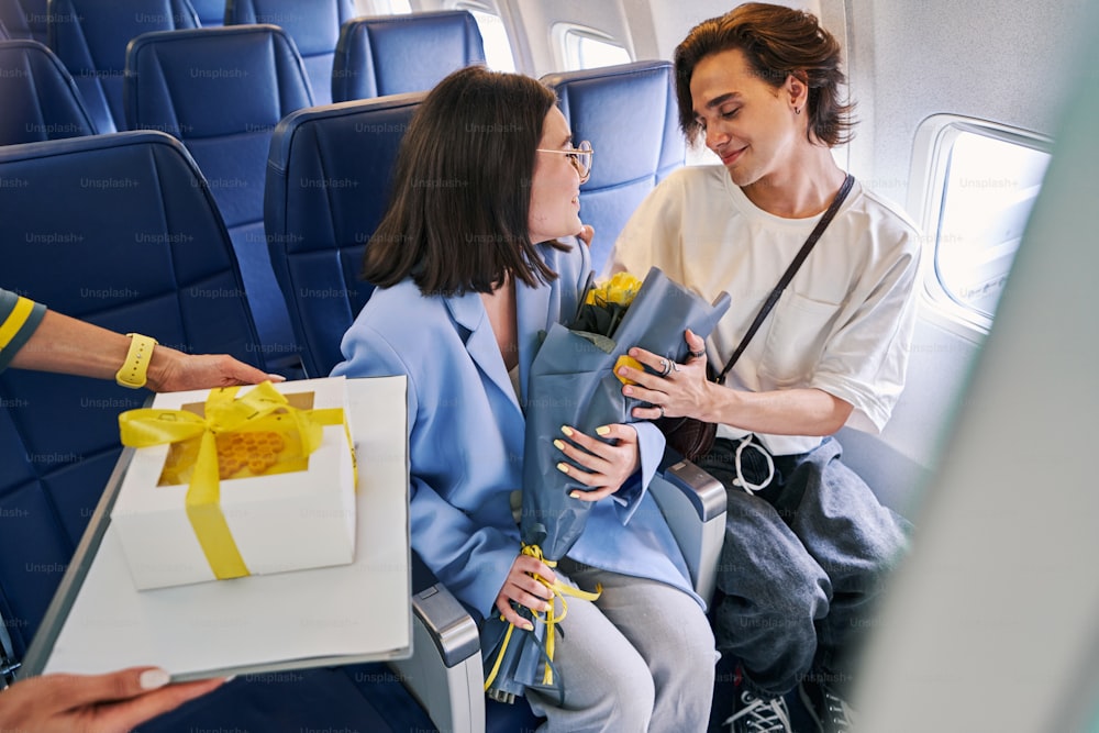 Dark-haired young woman with a bouquet of beautiful flowers and her pleased attractive boyfriend sitting in the aircraft cabin