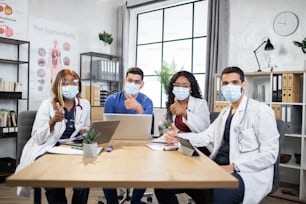 Confident male and female doctors in medical clothes and masks sitting at table with modern gadgets, and showing thumbs up on camera. Multiracial specialists on consilium. Approval concept.