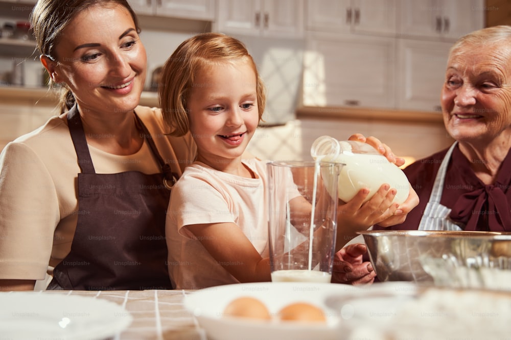 Delighted girl enjoying the process of pouring milk into big plastic cup on table while her family watching her