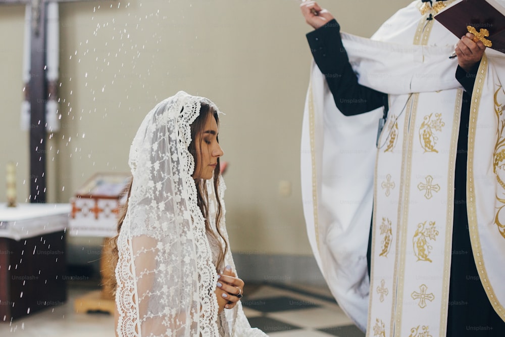 Priest blessing with holy water stylish bride in kerchief at altar during holy matrimony in church. Wedding ceremony in cathedral. Classic spiritual wedding bride praying