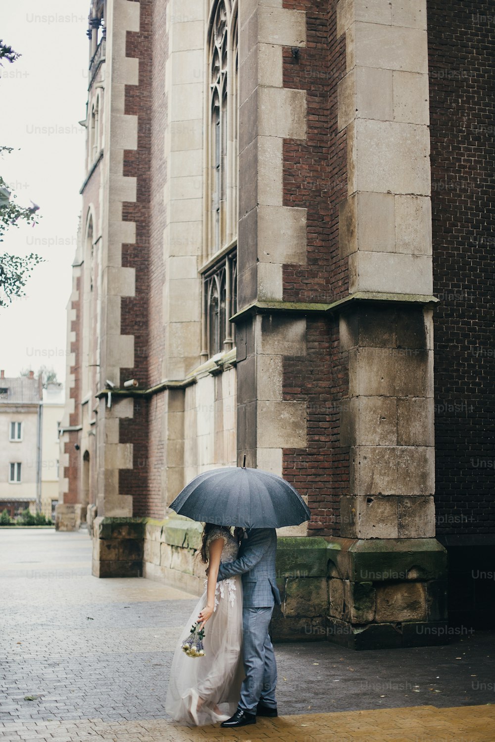 Stilvolle Braut und Bräutigam küssen sich unter dem Regenschirm vor dem Hintergrund der alten Kirche im Regen. Provence Hochzeit. Schönes Hochzeitspaar umarmt sich unter schwarzem Regenschirm in regnerischer Straße. Romantischer Moment