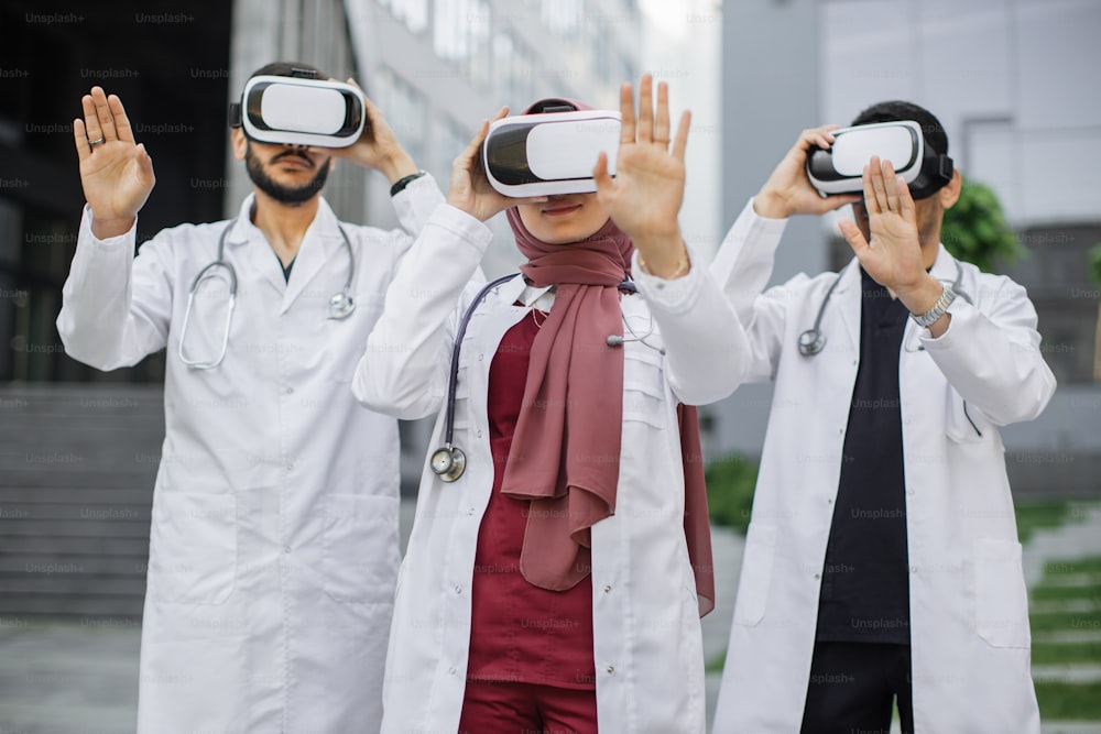 Outdoors shot of multiracial team of three male and female doctors wearing virtual reality glasses, train for better diagnostics, standing in front of modern clinic. Patient examination