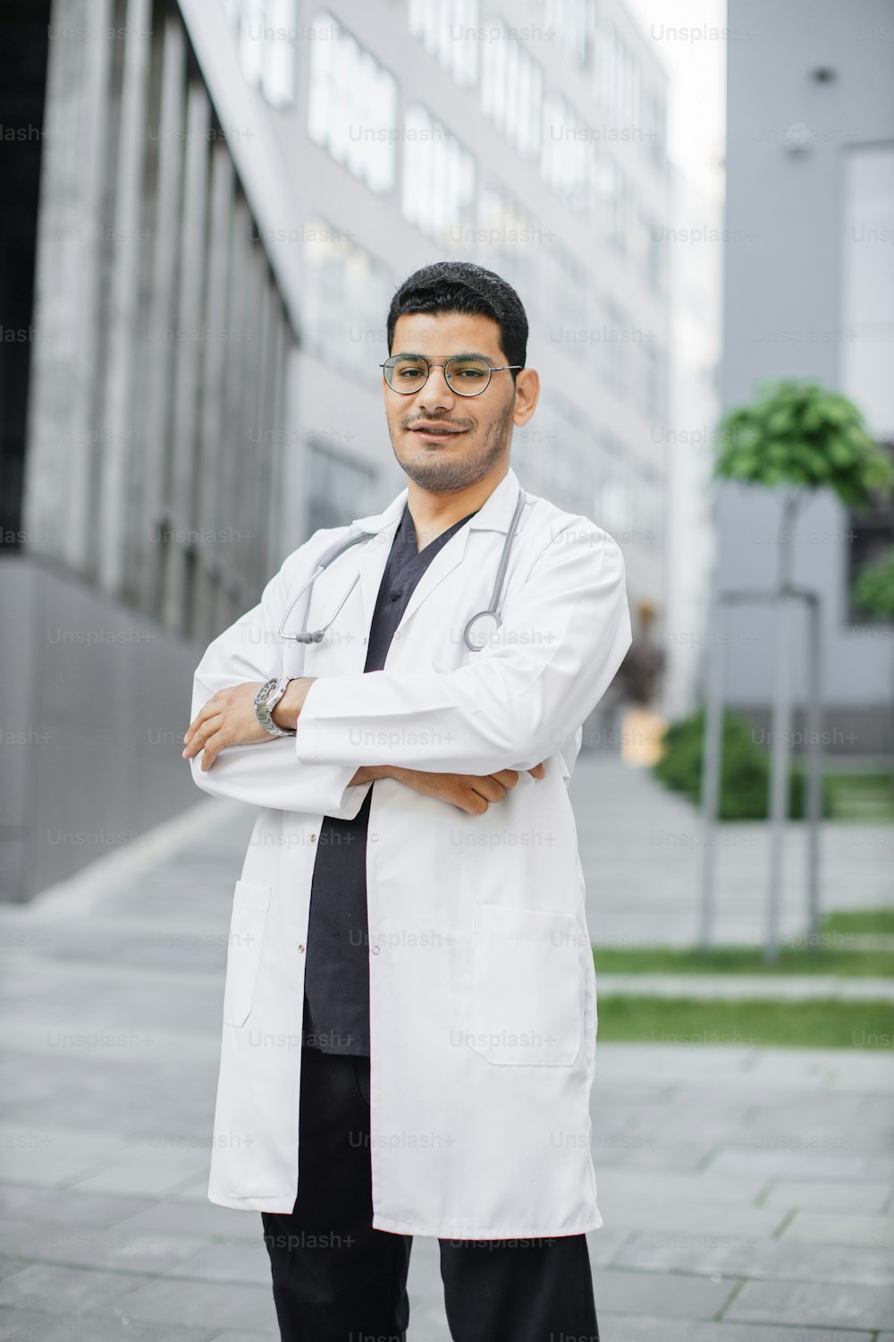 Portrait of young confident Asian Indian medical doctor standing outside hospital building with arms crossed and stethoscope around neck. Healthcare professional portrait