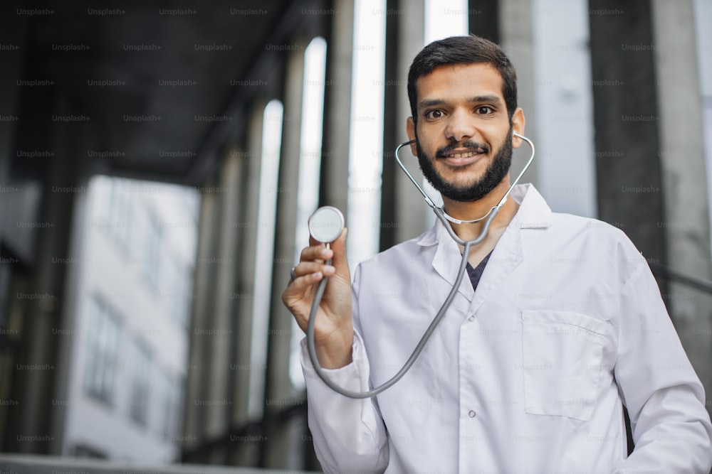 Portrait of smiling friendly male Arabian doctor or medical student in white coat, standing outside modern hospital, demonstrating his stethoscope to camera. Focus on face. Panorama, copy space