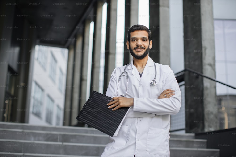 Sympathischer selbstbewusster arabischer männlicher Arzt im weißen Kittel und mit Stethoskop, Klemmbrett und Stift haltend, mit verschränkten Armen auf dem Hintergrund eines modernen Krankenhauses stehend.