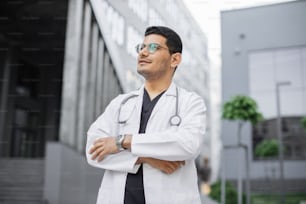 Ritratto ravvicinato di giovane medico arabo professionista maschio, che indossa camice bianco e stetoscopio intorno al collo, sognante che distoglie lo sguardo, in piedi all'aperto di fronte al moderno edificio ospedaliero