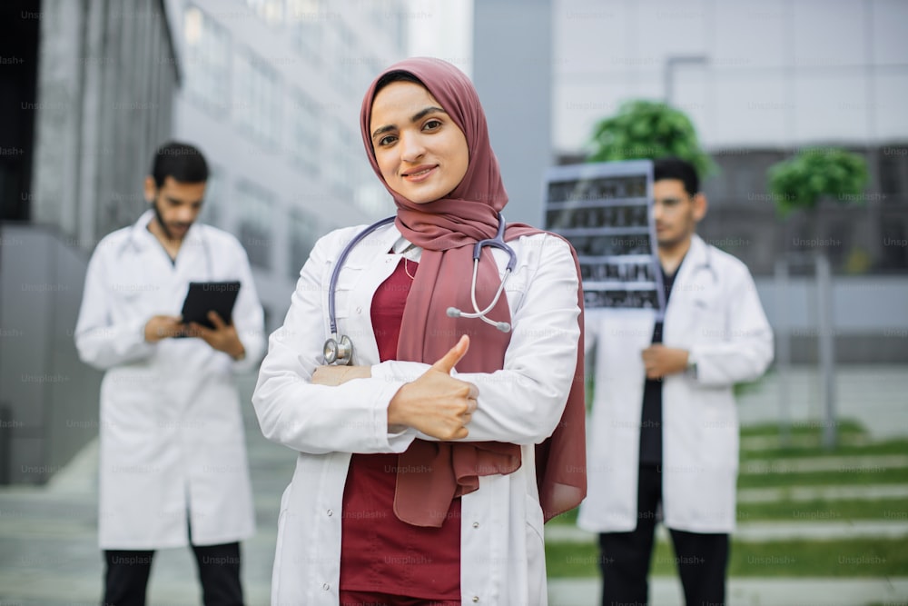Beautiful young female Arab doctor in uniform and hijab is looking at camera and smiling with thumb up while her two male confident colleagues are standing in background outdoors near modern hospital