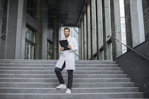 Vista frontale ravvicinata di un piacevole giovane medico indiano asiatico in camice bianco, in possesso di un blocco per appunti, in piedi sulle scale della clinica moderna all'aperto, sorridente e guardando la macchina fotografica. Inquadratura a figura intera