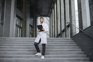 Close up front view of pleasant young Asian Indian doctor in white coat, holding a clipboard, standing on stairs of modern clinic outdoors, smiling and looking at camera. Full length shot