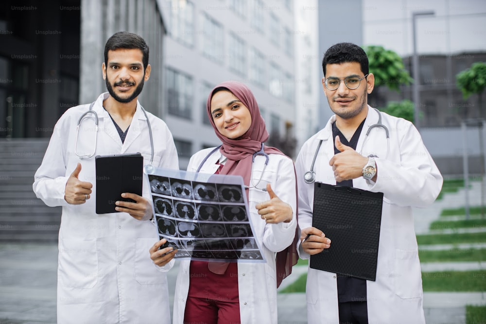 Portrait of three young Arabian doctors, posing to camera outdoors in front of modern clinic building, showing thumbs up. Radiologist doctors holding x-ray, clipboard and tablet pc.