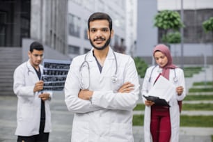Outdoor-Porträt eines selbstbewussten arabischen oder hinduistischen Arztes vor einem multiethnischen Team, der mit verschränkten Armen in die Kamera schaut und lächelt. Männliche und weibliche Kollegen, die am Hintergrund arbeiten.