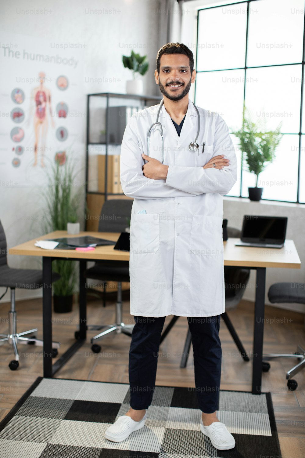 Smiling male Indian professional doctor in medical coat, standing with arms crossed, looking at camera. Happy confident Arab man general practitioner with stethoscope in office. Full length portrait