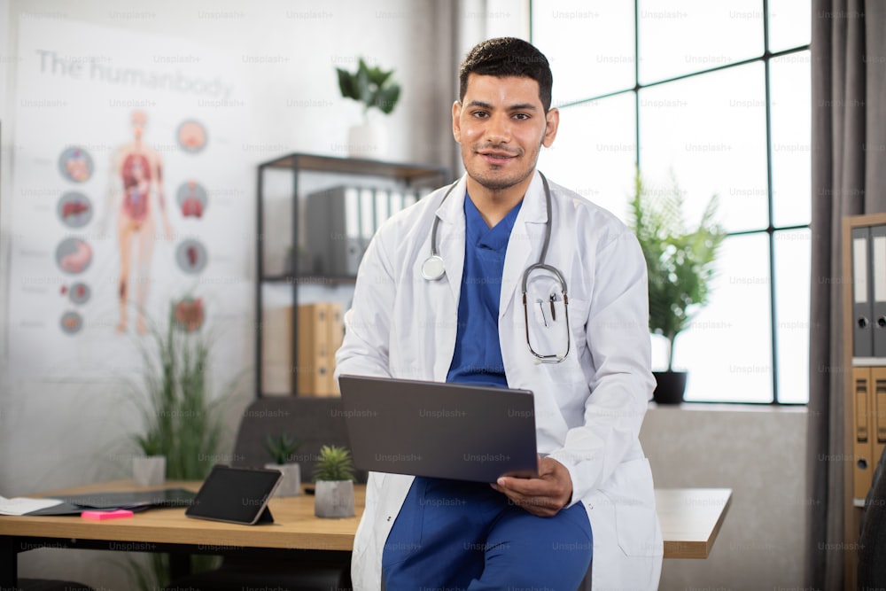 Portrait of pleasant young Arabian doctor in white coat and blue scrubs, with stethoscope around neck, leaning on the table in modern office and using laptop pc for medical records or prescribing