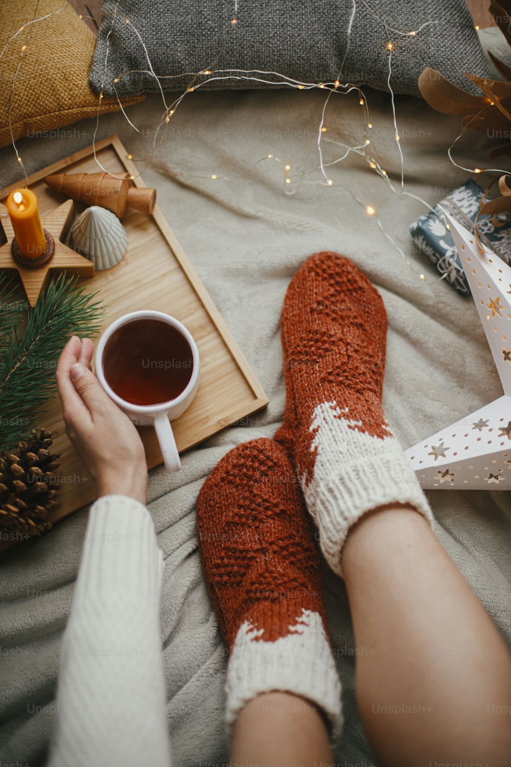 Jambes de femme dans des chaussettes chaudes sur une couverture douce avec une tasse de thé, des étoiles de Noël, des lumières dorées, des arbres, une bougie et des oreillers, vue de dessus. Des moments de détente agréables à la maison. Vacances d’hiver et d’automne.