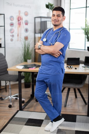 Handsome young doctor or medical student is looking at camera while standing with folded arms. Arabian male doctor posing with arms folded in modern light clinic office