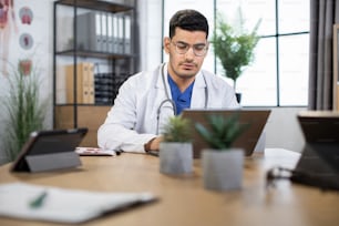 Professional medical specialist in uniform using wireless laptop for work at own cabinet. Confident focused skilled Arabian male doctor sitting at the table and typing on laptop