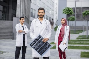 Confident team of multiracial three doctors in scrubs and coats, standing outdoors. Male Arabic doctor leader standing with tomography scan in hands and looking at camera with smile