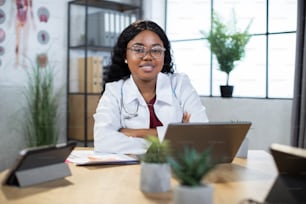 Close up of pretty smiling qualified black-skinned female doctor in white medical gown, sitting at the table in modern office with laptop computer and looking at camera