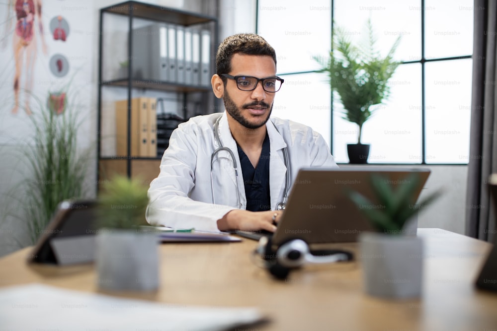 Indian arab male doctor typing on laptop, writing a prescription in his modern office. Young physician using telehealth medical chat for online appointment and consultation of patient on computer