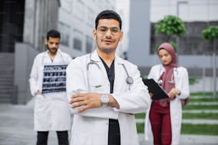 Outdoor portrait of confident Arabic or hindu man doctor in front of multiethnic team, looking at camera with arms crossed and smiling. Male and female colleagues working on background.