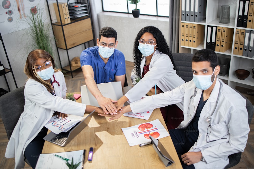 Success, teamwork concept. Team of multiracial scientists doctors in masks stacking hands together while sitting at table at modern hospital. Concept of international cooperation and health care.