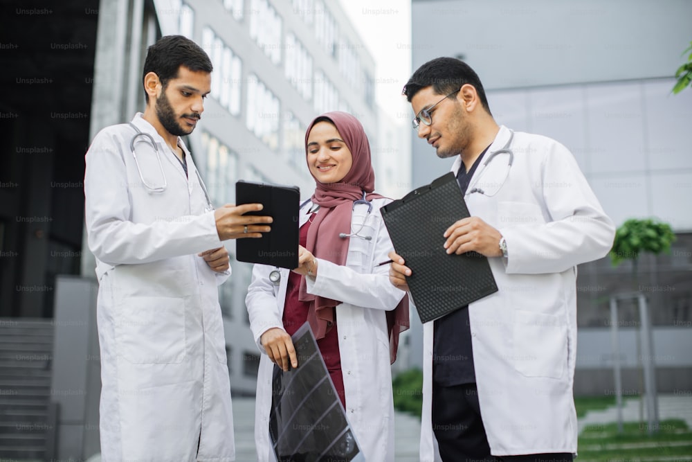 의료 유니폼을 입은 세 명의 젊은 다인종 의사, 히잡을 쓴 여성, 두 명의 남성이 태블릿 PC에서 CT 스캔 엑스레이를 보고 있습니다. 클리닉 앞에서 야외에서 토론하는 아라비아 의료진